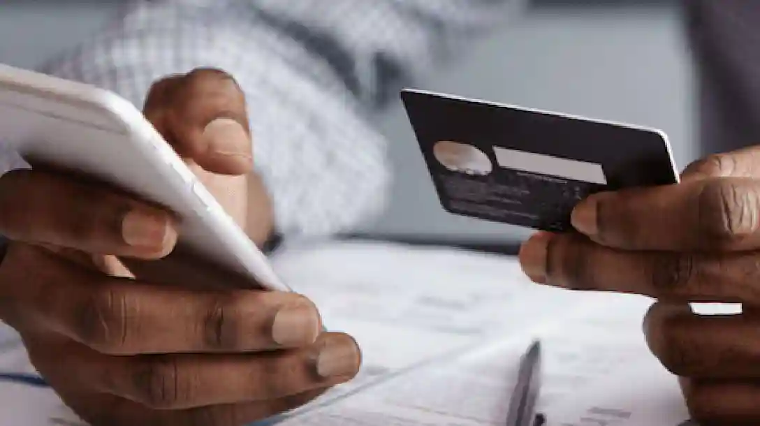 Person holding a phone in one hand and a credit card in the other, seated at a table with pen and paper on top.