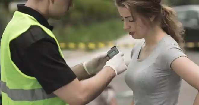 person in grey t shirt taking a breathalyzer test conducted by a police office wearing a neon yellow vest and medical gloves.