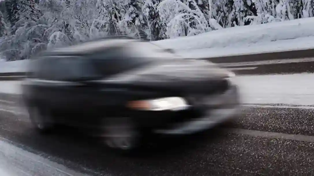 Black vehicle traveling down a road with snow capped landscape in the background.